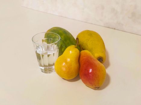 Glass of water and yellow fruits, papaya and pear on white bench in soft light.