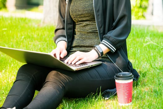 A woman sitting on the grass while using laptop computer with a red cup of coffee