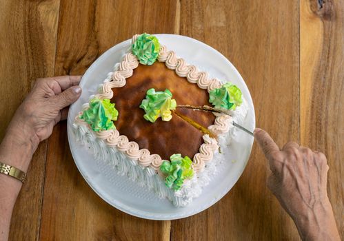 A cake on a table two wrinkled old hand, one cutting the cake and the other one holding the cake