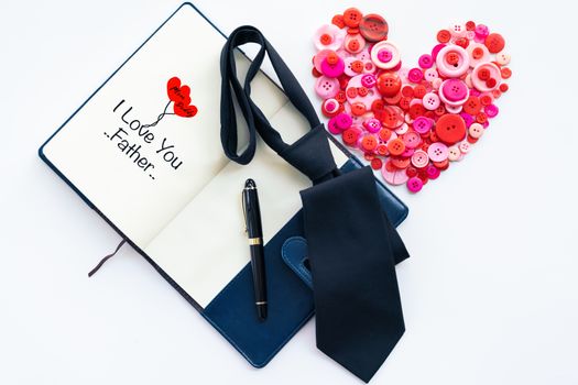 Happy fathers day, red button in heart shape and dark tie on book and luxury pen with text I Love you father