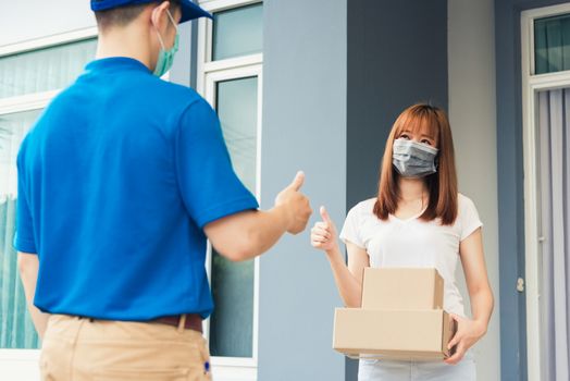 Asian delivery express courier young man giving parcel boxes to woman customer receiving both protective face mask and show thumbs up finger for good support sign, under curfew pandemic coronavirus