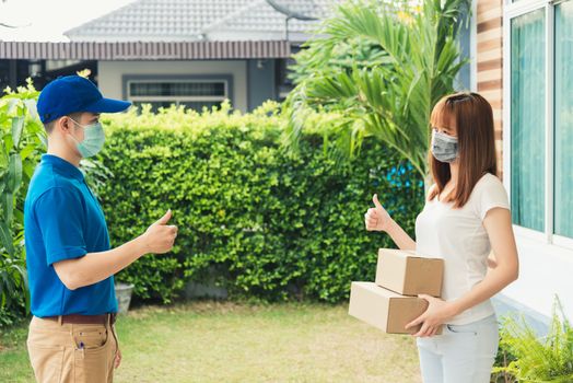 Asian delivery express courier young man giving parcel boxes to woman customer receiving both protective face mask and show thumbs up finger for good support sign, under curfew pandemic coronavirus