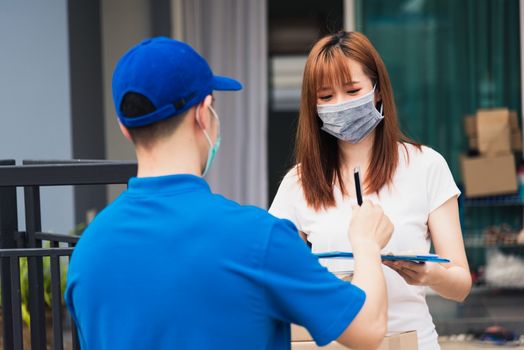 Asian young delivery man with package post boxed he protective face mask service woman customer using pen signature on paper book at front home, under curfew quarantine pandemic coronavirus COVID-19