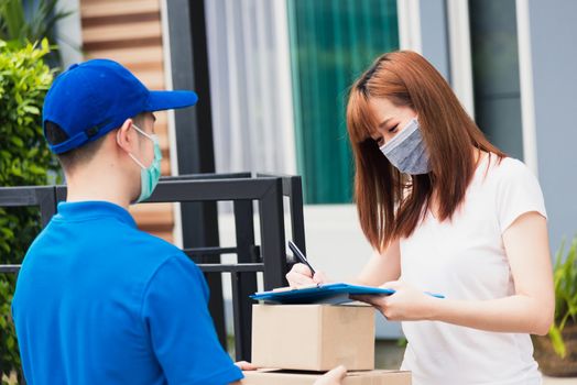 Asian young delivery man with package post boxed he protective face mask service woman customer using pen signature on paper book at front home, under curfew quarantine pandemic coronavirus COVID-19