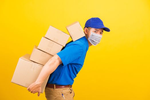Asian young delivery worker man in uniform wearing face mask protective he has many job lifting stack heavy a lot of boxes on back, under coronavirus COVID-19, studio shot isolated yellow background