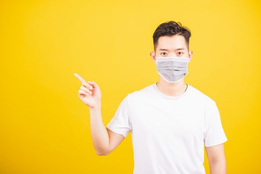 Portrait Asian young man wearing face mask protects filter dust pm2.5 anti-pollution, anti-smog, air pollution and COVID virus her pointing out side on yellow background, with copy space