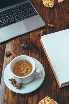 White cup of tea with star anise with a notebook on a wooden table with a computer in the background