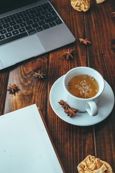 White cup of tea with star anise with a notebook on a wooden table with a computer in the background