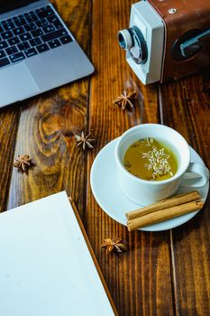 White cup of tea with cinnamon with a notebook on a wooden table with a computer in the background