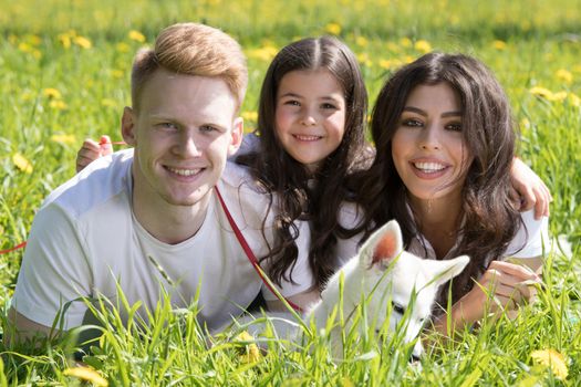 Happy family of parents and daughter laying on grass in park with pet husky puppy dog