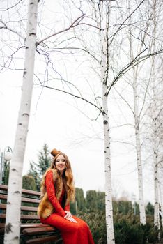 young girl with red hair in a bright red dress on a bench in an empty park among the birches