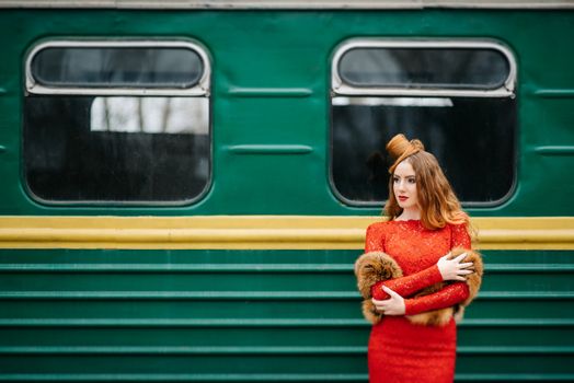 young girl with red hair in a bright red dress near an old green passenger car