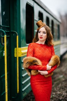 young girl with red hair in a bright red dress near an old green passenger car