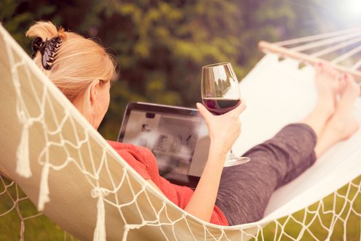 Beautiful woman relaxing on a hammock with glass of wine and browsing the web on a tablet (vintage effect).