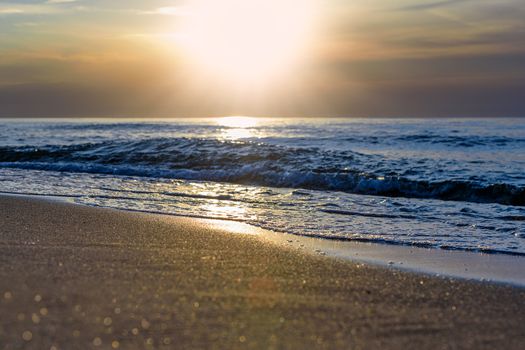 The setting sun is reflected in the waves of the sea and in the sand grains on the beach with a bokeh effect.