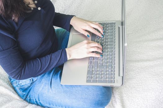 Young woman sitting on bed covered with white woolen blankets, with laptop on his lap. Home office work concept. Girl in jeans with a blue top and lacquered nails.