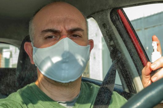 Turin, Piedmont, Italy. April 2020. Portrait of a Caucasian man driving the car wearing a white mask to avoid contagion. Mist alcohol to disinfect surfaces: droplets shine suspended in the air.