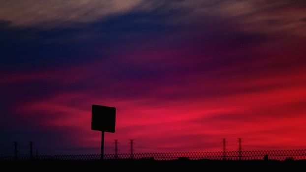 A road sign and electrical transmission towers form silhouettes against a vibrant pink, magenta and purple evening sky after sunset.