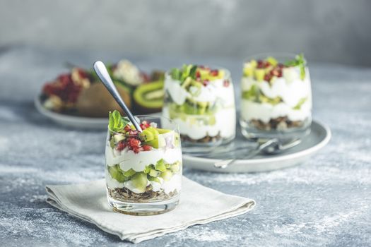 Glass jar of kiwi parfait dessert in glass with ingredients with spoon on gray napkin. Yogurt, granola and fruits. Healthy snack or breakfast. Light gray concrete surface.