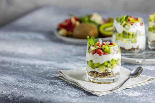Glass jar of kiwi parfait dessert in glass with ingredients and spoon on gray napkin. Yogurt, granola and fruits. Healthy snack or breakfast. Light gray concrete surface.