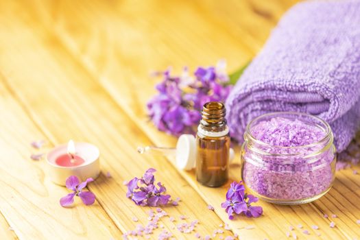 Spa still life with violet oil, towel, violaceous bath salt in glass jar and perfumed candle on natural wood table surface.