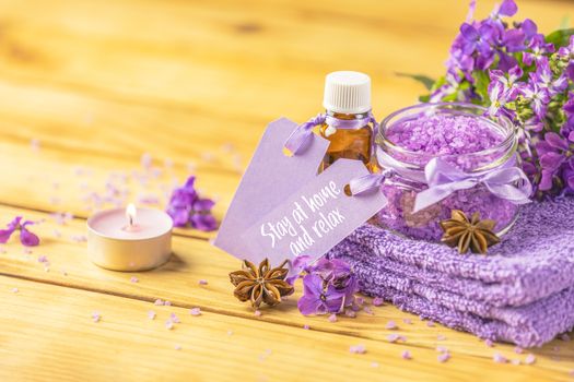 Stay at home and relax text phrase on label sticker. Spa still life with violet oil, towel and perfumed candle on natural wood table surface. Quarantine coronavirus life concept.