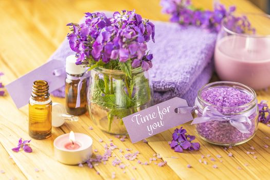 Time for you text phrase on label sticker. Spa still life with violet oil, towel, violaceous bath salt in glass jar and perfumed candle on natural wood table surface.