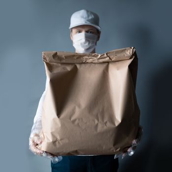 Safe food or goods delivery. Young courier delivering grocery brown eco paper bag order to the home of customer with mask and gloves during the coronavirus pandemic. White clothes gray background copy space for text