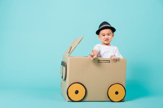 Happy Asian children boy smile in driving play car creative by a cardboard box imagination, summer holiday travel concept, studio shot on blue background with copy space for text