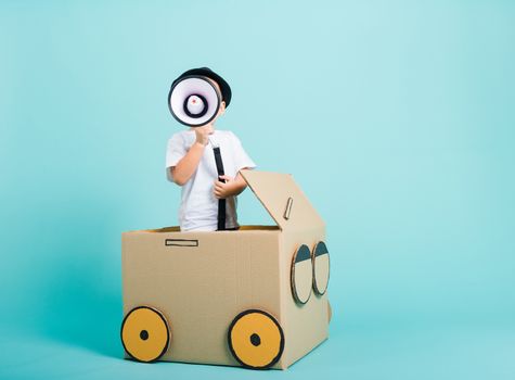 Happy Asian children boy smile in driving play car creative by a cardboard box imagination with megaphone, summer holiday travel concept, studio shot on blue background with copy space for text