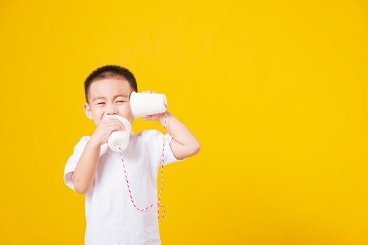 Portrait happy Asian cute little children boy smile standing so happy wearing white T-shirt playing paper can telephone, studio shot on yellow background with copy space