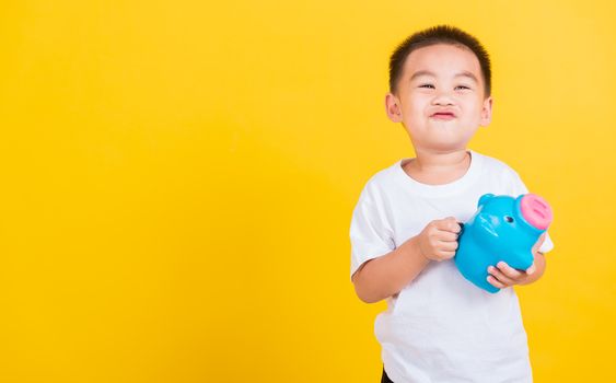 Asian Thai happy portrait cute little cheerful child boy smile putting coin money to the piggy bank and looking camera, studio shot isolated on yellow background with copy space