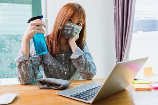 Asian Business young woman work from home office he cleaning laptop computer by sanitizer alcohol spray quarantines disease coronavirus or COVID-19 and wearing a protective mask before do job