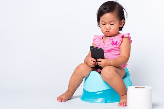 Asian little cute baby child girl education training to sitting on blue chamber pot or potty and play smart mobile phone with toilet paper rolls, studio shot isolated on white background, wc toilet