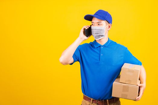 Asian young delivery worker man t-shirt and cap uniform wearing face mask protective use smart mobile phone call to customer under coronavirus or COVID-19, studio shot isolated yellow background