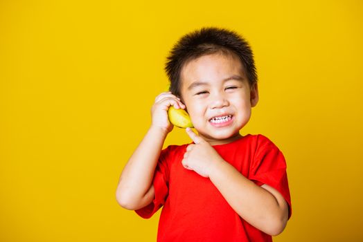 Happy portrait Asian child or kid cute little boy attractive smile wearing red t-shirt playing holds banana fruit pretending to be like a telephone, studio shot isolated on yellow background