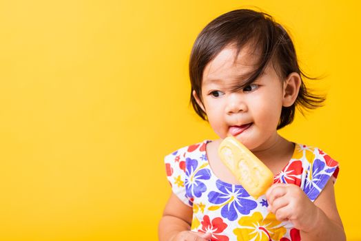 Happy portrait Asian baby or kid cute little girl attractive laugh smile wearing dick pattern shirt holds and eating sweet wooden ice cream, studio shot isolated on yellow background, summer concept