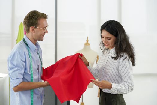 Fashion designer room, man and woman working in home base cloth design business. Men's dressmaker presents red fine fabrics to young customers.