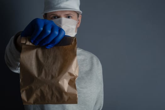 Safe food or goods delivery. Young courier delivering small brown eco paper bag order to the home of customer with mask and gloves during the coronavirus pandemic. Gray background copy space for text