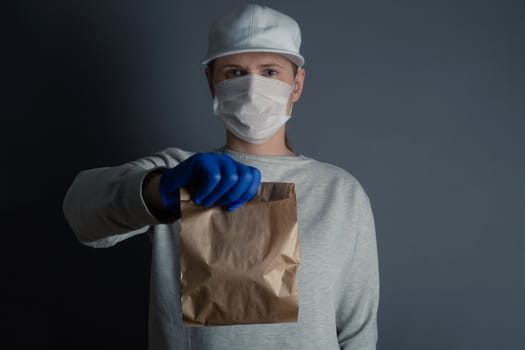 Safe food or goods delivery. Young courier delivering small brown eco paper bag order to the home of customer with mask and gloves during the coronavirus pandemic. Gray background copy space for text