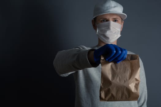 Safe food or goods delivery. Young courier delivering small brown eco paper bag order to the home of customer with mask and gloves during the coronavirus pandemic. Gray background copy space for text
