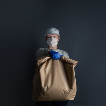 Safe food or goods delivery. Young courier delivering grocery big brown eco paper bag order to the home of customer with mask and gloves during the coronavirus pandemic. Gray background copy space for text