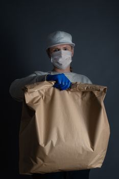 Safe food or goods delivery. Young courier delivering grocery big brown eco paper bag order to the home of customer with mask and gloves during the coronavirus pandemic. Gray background copy space for text