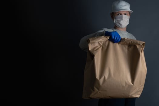 Safe food or goods delivery. Young courier delivering grocery big brown eco paper bag order to the home of customer with mask and gloves during the coronavirus pandemic. Gray background copy space for text