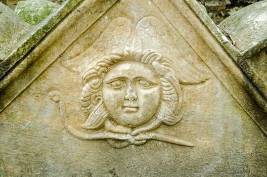 An ancient carving of an angel with a chubby face and what looks like a snake around its neck and wings behind.  Stone sarcophagus dating from late Roman times and left on the street outside the archaeology museum in Istanbul, Turkey.  