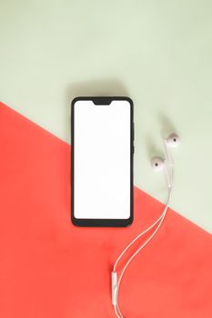 Modern smartphone with white screen and headphones on vivid background. Top view of a phone and headset against red and green background