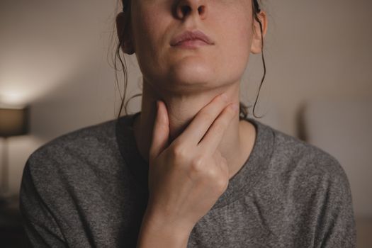 Sore throat, virus disease or flu. Woman holds hand against her throat feeling pain in bedroom