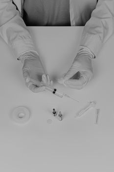 MD preparing injection, faded monochrome style. Top view of medical doctor hands in gloves and lab coat holding syringe on white table with medical accessories