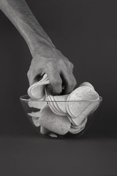 Grabbing potato chips from a glass bowl. Male hand greedily takes handful of crisps out in black monochrome background