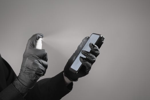 Cleaning the phone with a sanitizer, total black concept. Hands in black gloves spray sanitizer on the smartphone screen. Virus spread prevention concept